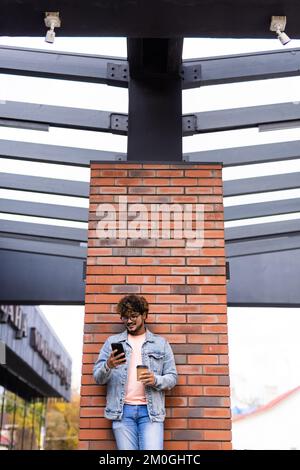 Lächelnder Indianer, der sein Handy benutzt, SMS liest, online im Freien kommuniziert. Stilvoller, fröhlicher asiatischer Hipster, der eine Tasse Kaffee hält Stockfoto