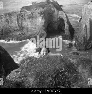 1950er, historisch, an der Küste, hoch über dem Ozean, mit Blick auf das Meer und hervorstehende Felsen, eine Dame unter der Erde, die vorgibt, sich an der Klippe oder dem Überhang zu Klammern, England, Großbritannien. Stockfoto