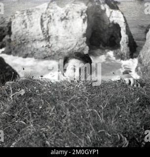 1950er, historisch, an der Küste, hoch über dem Ozean, eine Dame, die ihr halbes Gesicht unter der Erde versteckt und so tut, als würde sie sich an der Klippe festhalten oder überhängen, um einen Freund, England, zu überraschen. Stockfoto