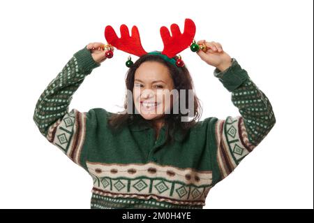 Wunderschöne Frau trägt einen grünen Pullover mit Xmas-Muster, hält glänzende Bälle auf dem Reifen mit Hirschgeweih, lächelt vor der Kamera Stockfoto