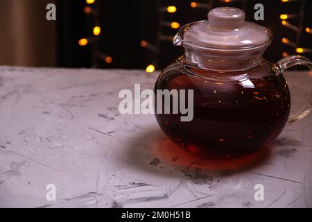 Wasserkocher aus Fotoglas mit Tee, der auf dem Tisch steht, mit einer Weihnachtsgirlande im Hintergrund Stockfoto