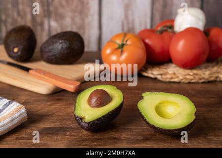 Avocado in zwei Stücke geschnitten, mit Holzhintergrund mit Tomaten, Knoblauch und mehr Avocados. Stockfoto