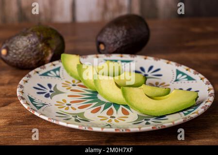 Avocado-Stücke auf einem rustikalen Teller auf einem Holztisch, begleitet von weiteren Avocados. Stockfoto