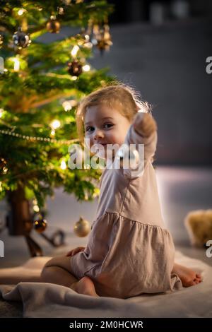 Blick von hinten, ein süßes kleines Mädchen, das zu Hause Bälle an den Weihnachtsbaum hängt. Stockfoto