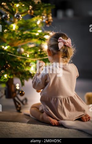 Blick von hinten, ein süßes kleines Mädchen, das zu Hause Bälle an den Weihnachtsbaum hängt. Stockfoto