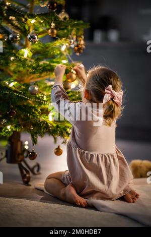 Blick von hinten, ein süßes kleines Mädchen, das zu Hause Bälle an den Weihnachtsbaum hängt. Stockfoto