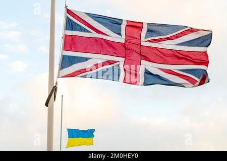 Union Jack und ukrainische Flagge, die mit Halbmast gegen einen stumpfen, blassblauen und weißen Himmel anlässlich des Todes von Königin Elizabeth II. Fliegen Stockfoto