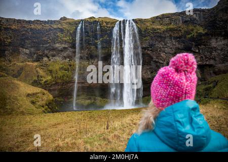 Island, Sudurland, eine Figur mit blauer Jacke und einer rosa Kappe von hinten im Vordergrund, blickt auf den Seljalandsfoss Wasserfall, 60 m hoch, einer der wenigen Wasserfälle, hinter denen Sie vorbeifahren können Stockfoto
