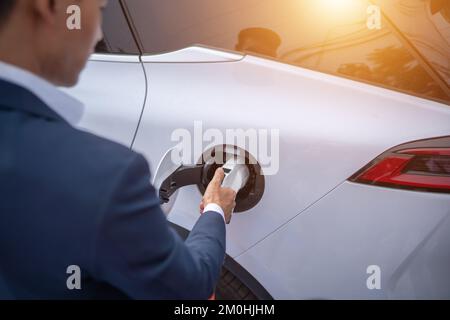 Ein junger Geschäftsmann lädt ein Elektrofahrzeug an einer Ladestation für Elektrofahrzeuge auf. Stockfoto