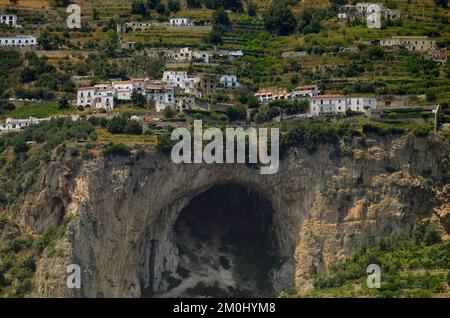 Eine große Höhlenöffnung befindet sich an der Amalfiküste inmitten der Kalksteinklippen mit Häusern, die auf unheimlich hohen Häusern mit Hotel und Strand unten stehen. Stockfoto