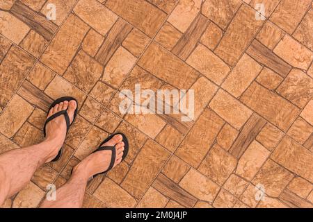 Die Beine eines Mannes in schwarzen Flip-Flops stehen auf einem braunen Fliesenboden mit einem abstrakten Steinmuster im Hintergrund, Draufsicht. Stockfoto