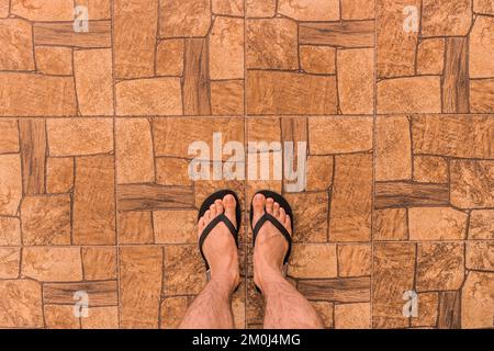 Die Beine eines Mannes in Flip-Flops stehen auf einem braunen Fliesenboden mit abstraktem Steinmuster im Hintergrund, Draufsicht. Stockfoto