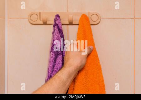 Die Hand des Mannes nimmt ein orangefarbenes Handtuch neben einem violetten Handtuch im Badezimmer. Pflege- und Hygienekonzept. Stockfoto