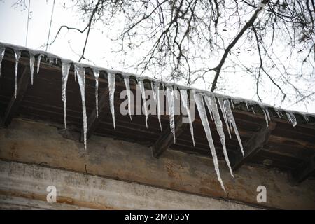 Große Eiszapfen unter dem Dach eines gelben Hauses an einem sonnigen, klaren Tag. Blauer Himmel im Hintergrund Stockfoto