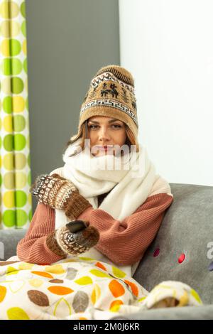 Eine junge Frau, die auf dem Sofa sitzt, einen Wollmütze, einen Schal und Handschuhe trägt und eine Fernbedienung für Sky tv in der Hand hält Stockfoto