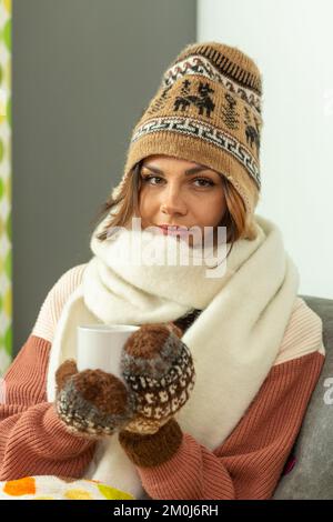 Eine junge Frau sitzt auf einem Sofa, trägt einen Wollmütze, einen Schal und Handschuhe drinnen und hält eine Kaffeetasse. Stockfoto
