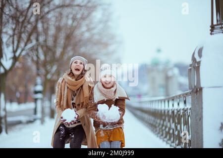 Lächelnde, stilvolle Mutter und Tochter im Mantel, Hut, Schal und Fäustlingen werfen im Winter im Freien in der Stadt Schnee. Stockfoto