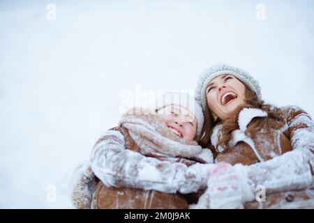 Lächelnde stilvolle Mutter und Kind im Mantel, Hut, Schal und Fäustlinge liegen im Winter im Stadtpark im Schnee. Stockfoto