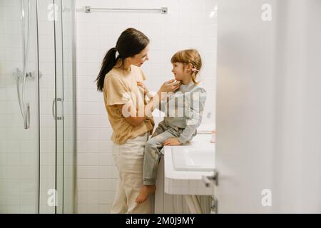 Weiße Frau lächelt, während sie mit ihrer Tochter im Badezimmer zu Hause spricht Stockfoto