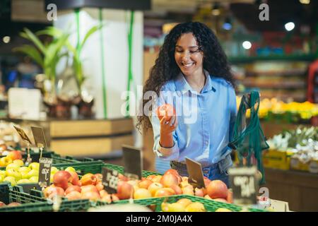 Eine hübsche junge Hispanic-Frau kauft in einem Supermarkt ein. Sie steht lächelnd in der Gemüse- und Fruchtabteilung, pflückt und hält einen Apfel in der Hand, steckt ihn in einen Öko-Sack. Stockfoto