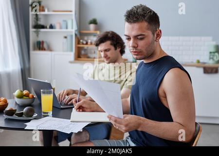 Porträt eines jungen schwulen Paares, das zusammenlebt, mit Fokus auf den Mann, der am Küchentisch lernt Stockfoto