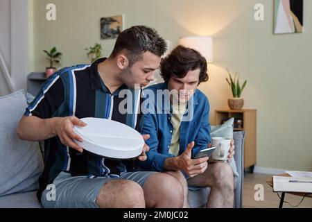 Porträt von zwei jungen Männern, die einen drahtlosen Roboter-Staubsauger über ein Smartphone in einem modernen Smart-Home einrichten Stockfoto