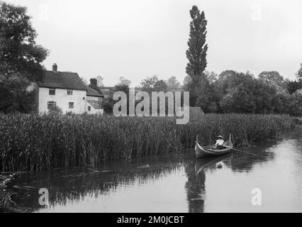 Schwarzweißes englisches Foto aus dem späten 19.. Jahrhundert, das eine Frau in einem Ruderboot auf einem Fluss zeigt, neben einigen großen Anlässen mit einem Haus oder einer Hütte in der Nähe. Stockfoto
