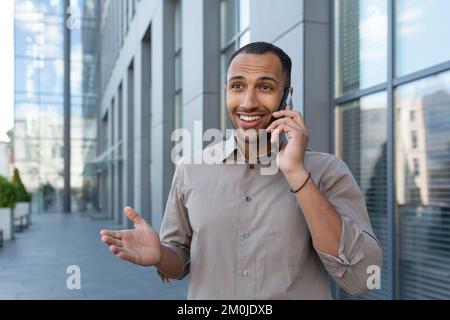 Fröhlicher und lächelnder afroamerikanischer Büroangestellter, der am Telefon telefoniert, ein Mann vor dem Bürogebäude im Hemd, der in der Stadt spaziert, ein Geschäftsmann in der Mittagspause. Stockfoto