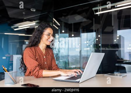Glückliche und lächelnde hispanische Geschäftsfrau, die auf dem Laptop tippt, Büroangestellte mit lockigem Haar und Brille, die mit ihren Ergebnissen zufrieden ist, bei der Arbeit im Bürogebäude. Stockfoto