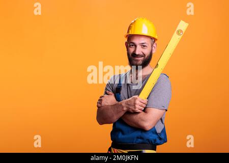 Ich repariere das Wasserstandlineal im Studio, stehe mit gekreuzten Armen. Junger Mechaniker, der sich auf Messungen mit einem professionellen Nivellierwerkzeug vorbereitet, selbstbewusster Tischler. Stockfoto