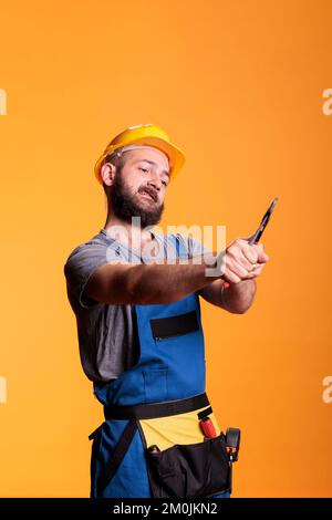 Bauunternehmer, der zur Reparatur eine Zange verwendet, als Zimmermann oder Konstrukteur im Studio arbeitet. Professioneller Bauarbeiter mit Hutzange und Renovierungswerkzeugen im Hintergrund. Stockfoto