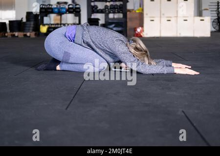 Seitenansicht einer blonden, erwachsenen Frau, die sich während ihres Vinyasa-Flow-Yoga-Trainings allein in der Kinderposition ausruht und Sportbekleidung im Fitnessstudio trägt. Stockfoto
