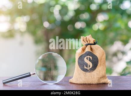 Budget- und Sparkonzept. Vergrößerungsglas neben einer Tasche voller Geld auf einem unscharfen Hintergrund. Stockfoto