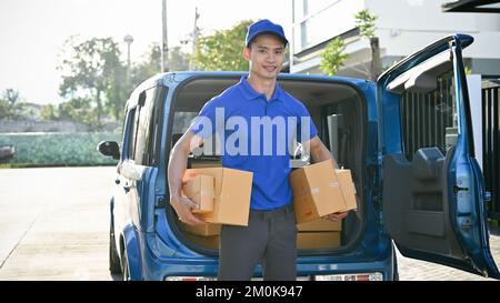 Junger asiatischer Liefermann, der Pakete mit sich führt und vor seinem Auto steht, während des Vertriebs Stockfoto
