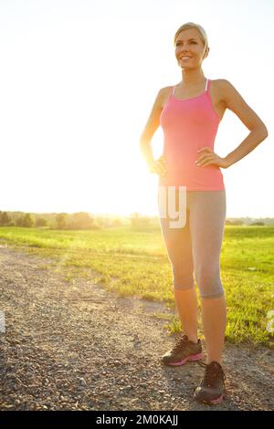 Genießen Sie die Landschaft, während Sie sich entspannen. Eine junge Frau in Sportbekleidung, die auf einer unbefestigten Straße neben einem Feld steht. Stockfoto