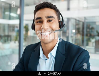 Porträt eines jungen, glücklichen, gemischten Rassen männlichen Callcenter-Agenten, der Anrufe entgegennimmt, während er bei der Arbeit ein Headset trägt. Ein hispanischer Sekretär lächelt Stockfoto