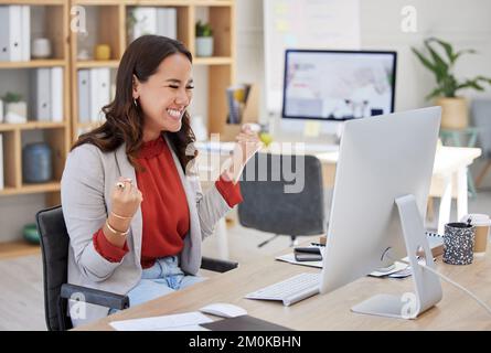 Eine asiatische Geschäftsfrau gemischter Rassen, die Freude ausdrückt und einen Sieg in ihrem Bürojob feiert. Eine Chinesin, die erfolgreich aussieht, während sie einen Desktop verwendet Stockfoto
