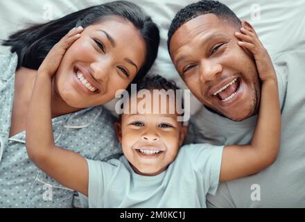 Porträt eines entzückenden kleinen Jungen mit den Händen seiner Eltern, die sie auf einem Bett liegen und sie in die Nähe ziehen. Netter Sohn, der zwischen seiner Mutter und liegt Stockfoto