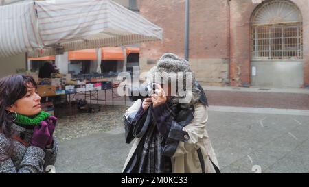 Junge Erwachsene hispanische Frau, die im Winter allein in einer Bar im Freien sitzt und grüne Handschuhe mit Polo-V-Ausschnitt trägt, schwere graue Jacke, fotografiert von weiblichem Senior Pho Stockfoto