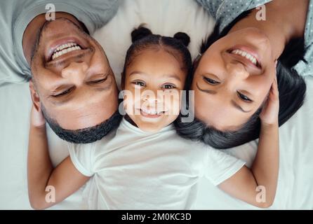 Entzückendes kleines Mädchen, das ihre Eltern nahe zieht, während es zwischen Mutter und Vater liegt, von oben. Gesichter liebevoller Eltern, die sich mit ihren verbinden Stockfoto