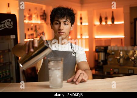 Der junge Barkeeper schüttet den fertigen Cocktail aus dem Shaker in das Glas. Barkeeper in grauer Schürze im Hintergrund. Speicherplatz kopieren Stockfoto