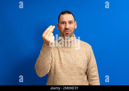 Ein gutaussehender lateinamerikanischer Mann mit Bart trägt einen braunen Rollkragenpullover auf blauem, isoliertem Hintergrund und macht selbstbewusst italienische Gesten Stockfoto