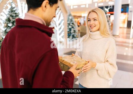 Rückblick auf einen unbekannten Mann, der an Heiligabend eine Geschenkbox mit Geschenk für eine bezaubernde blonde Frau in der Halle des Celebrate Shopping Mall hält Stockfoto