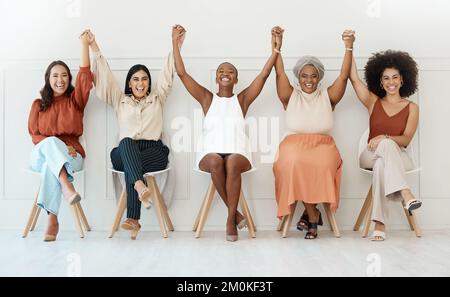 Eine Gruppe von fünf glücklichen jungen Geschäftsfrauen, die in einem Büro an einer Wand sitzen und gemeinsam die Hand halten. Lächelnde Geschäftsleute, die Händchen hielten und Stockfoto