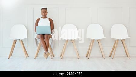 Junge fokussierte afroamerikanische Geschäftsfrau, die an einem Laptop arbeitet, während sie auf das Interview wartet, sitzt auf einem Stuhl an einer weißen Wand. Warten auf ein Stockfoto