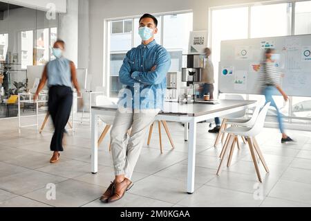 Junger asiatischer Geschäftsmann, der mit gekreuzten Armen steht und eine Maske bei der Arbeit trägt. Ein selbstbewusster chinesischer männlicher Boss trägt eine Maske, die vor A schützt Stockfoto