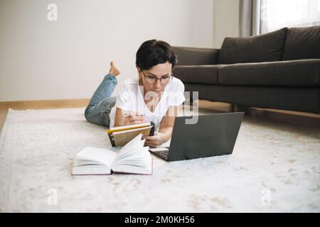 Frau arbeitet von zu Hause aus mit einem Laptop. Attraktive Brünette mittleren Alters mit Computer auf dem Teppich Stockfoto