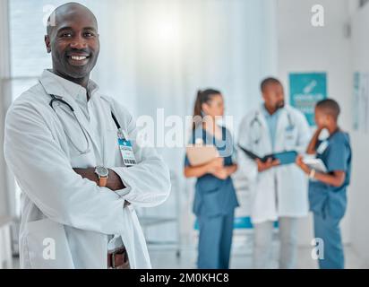 Reifer afroamerikanischer Arzt, der während der Arbeit in einem Krankenhaus mit gekreuzten Armen steht. Ein erfahrener Arzt lächelt im Stehen Stockfoto