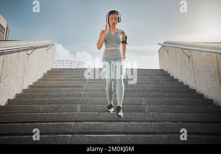 Junge gemischte Sportlerin hört Musik über Kopfhörer, während sie draußen die Treppen eines Gebäudes hinunterläuft. Junge Frau konzentrierte sich auf ihre Geschwindigkeit Stockfoto