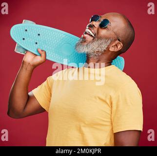 Ein reifer afroamerikanischer Mann, der mit einem Mini-Skateboard im Studio steht, isoliert vor rotem Hintergrund. Ein gutaussehender und sorgenfreier Mann Stockfoto